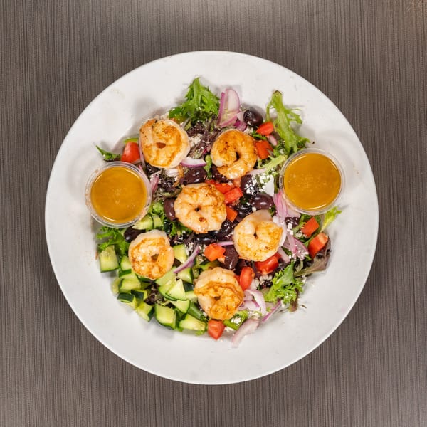 Overhead view of a shrimp salad with mixed greens, cucumbers, tomatoes, red onions, and two dressing cups.