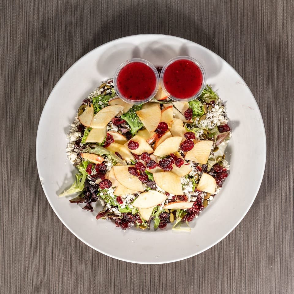 Overhead view of a vibrant salad with apples, cranberries, feta, and two cups of dressing.