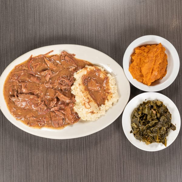 Pot roast with gravy, grits, collard greens, and sweet potato casserole on a dark table.